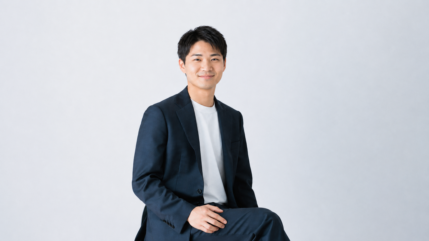 白い背景でジャケットを羽織り椅子に座る若手男性